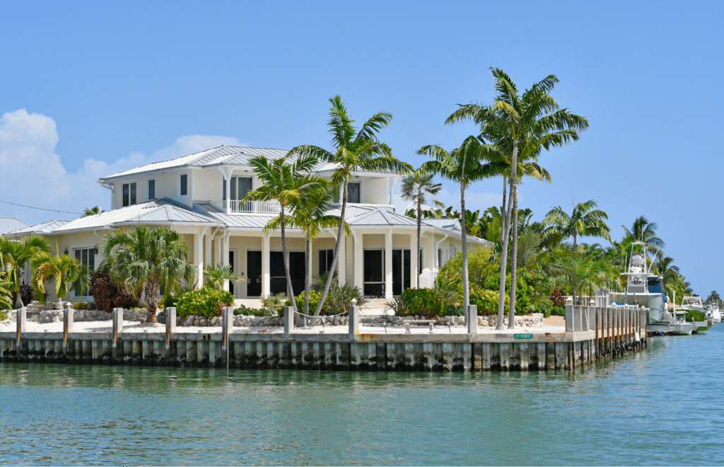 A luxury two-story waterfront estate with a metal roof and white columns, surrounded by tropical palm trees along a sea-walled canal with a docked yacht in Clearwater Beach, Florida.